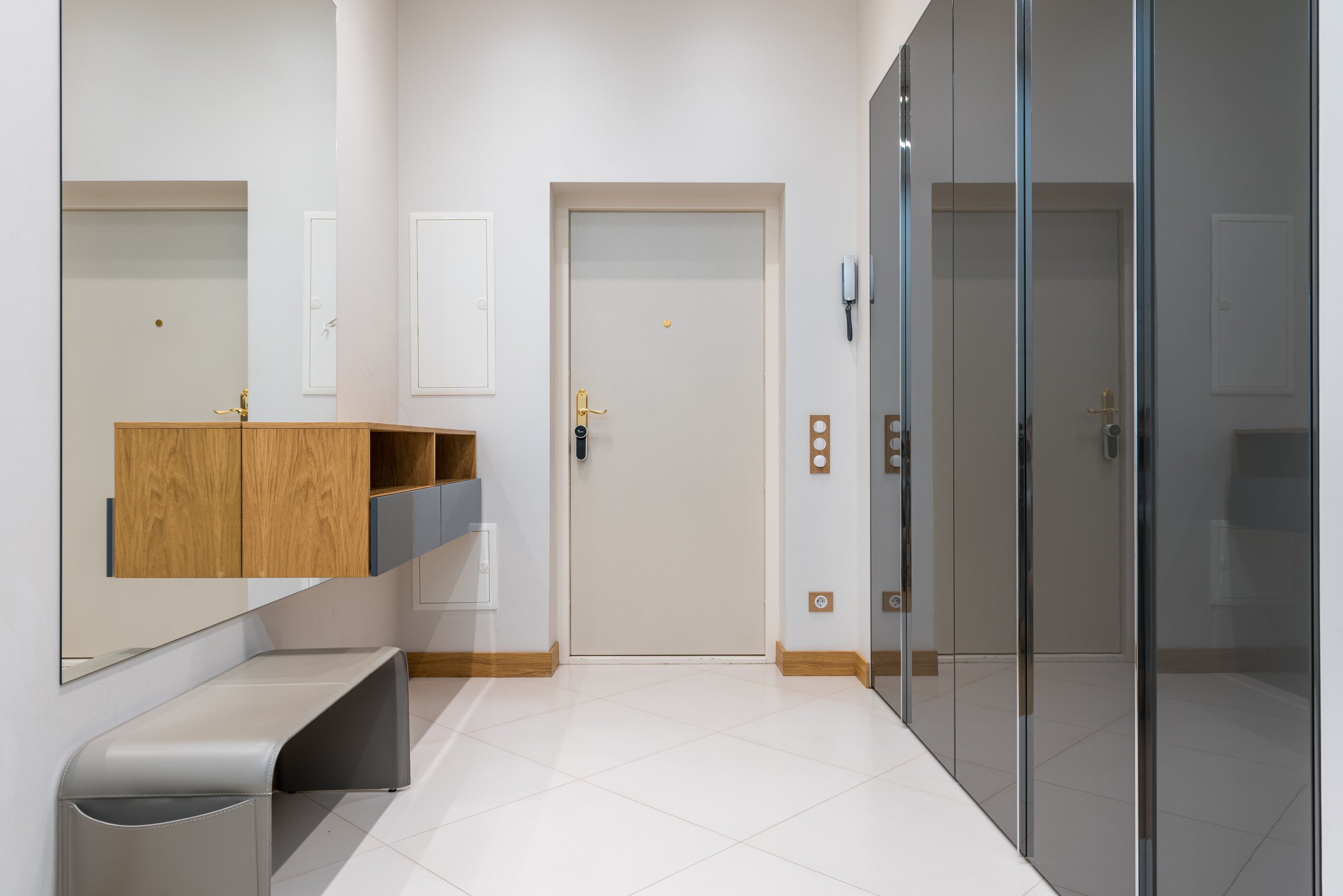 Presvetlená, minimalistická predsieň pozostávajúca z veľkej, lesklej, zasúvacej skrine sivej farby s leskom, dreveného botníku z masívneho dreva a minimalistickej lavice na sedenie v sivej farbe, nad ktorou je zrkadlo bez rámu. Lesklá podlaha a vchodové dvere sú bielej fabry