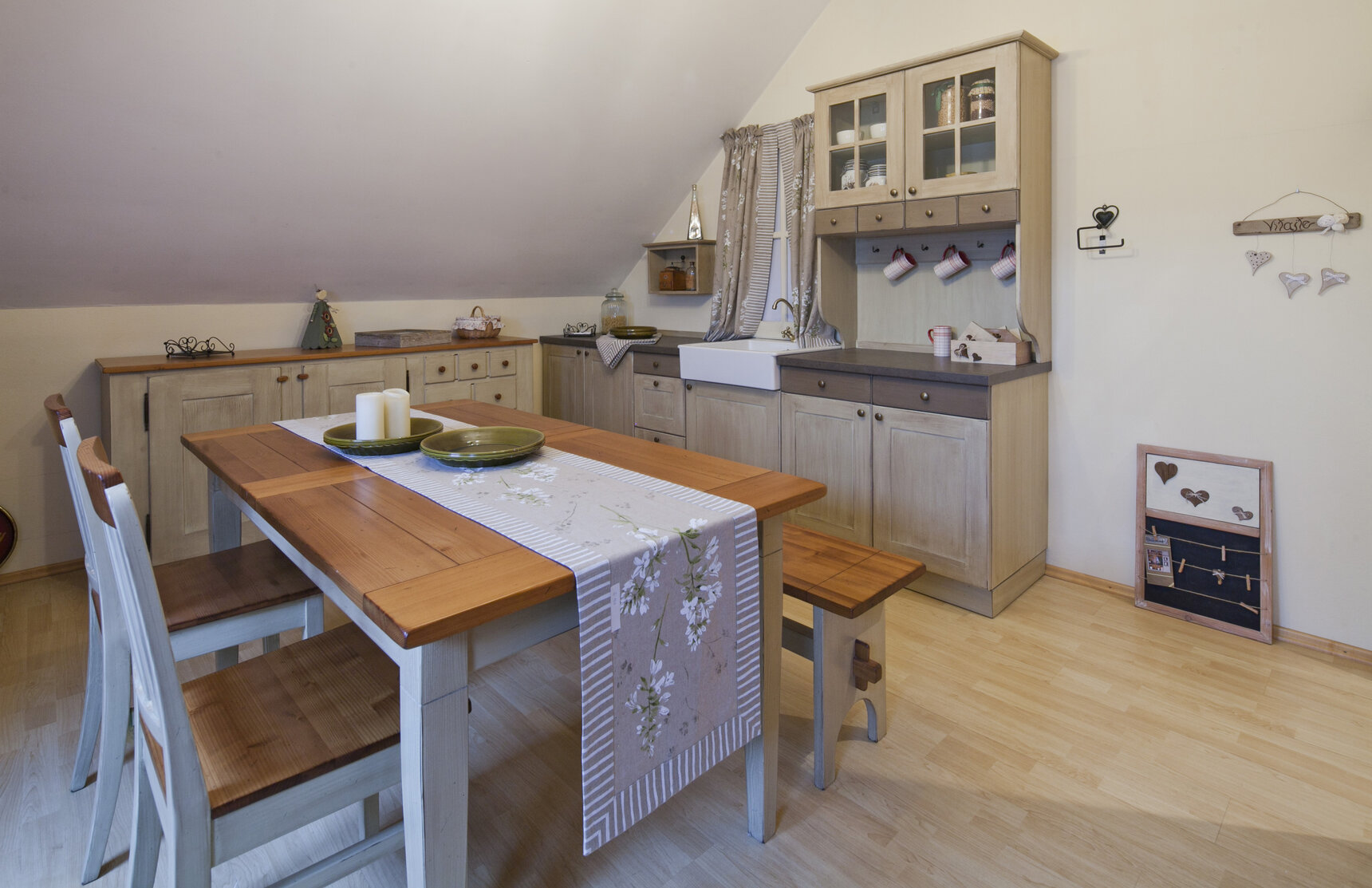 Rustikálna kuchyňa v svetlo hnedej farbe s jedálenským stolom, stoličkami a lavicou. Kuchynská doska je z dreva a dvierka na kuchynskej linke majú drevené okrúhle úchytky